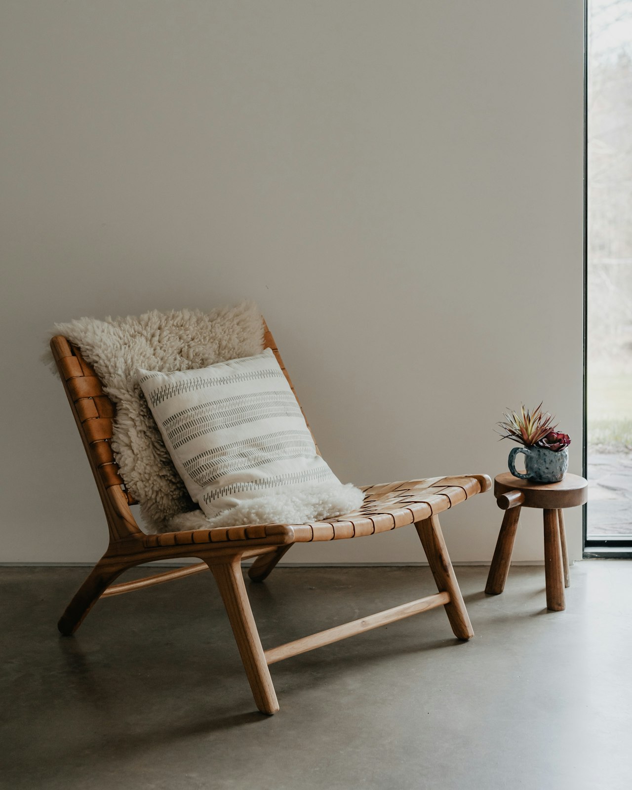 A woven oak lounge chair with a sheepskin and linen cushion on a concrete floor