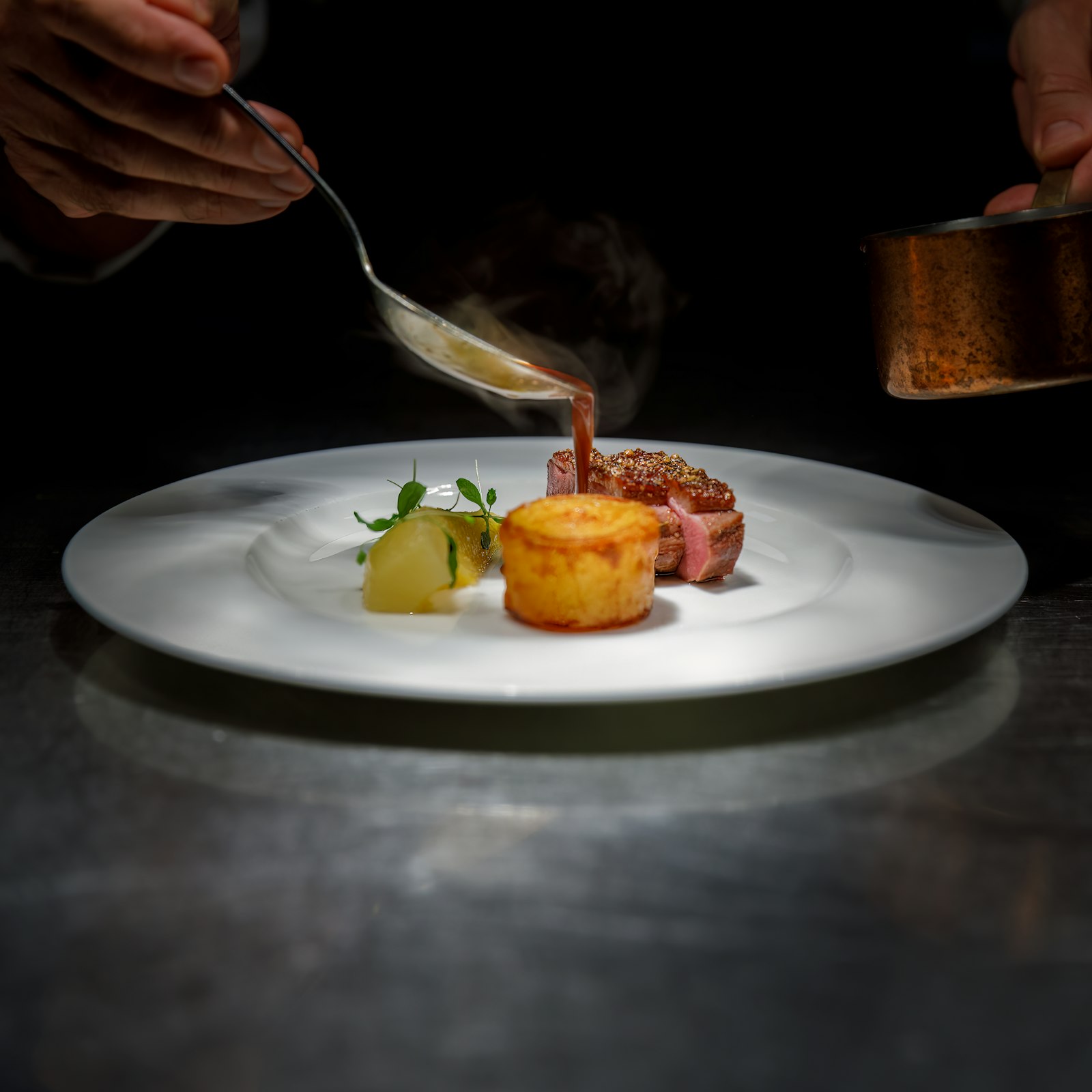 A chef spooning sauce onto a plated dish in a dark kitchen